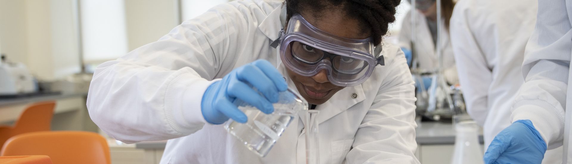 Post Bacc Student in lab gear pouring liquid into a beaker
